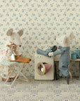 Two Maileg mouse figures—a dress-wearing one ironing and a pajama-clad one loading laundry—enjoy the Maileg Washing Machine in a cozy miniature room, complete with wardrobe, table, teapot, cups, and a floral patterned floor.