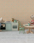 Two Maileg toy mice in striped shirts create a cosy dinner scene: one cooks at the stove, while the other sits at the Maileg Oval dining table. The miniature setting features patterned wallpaper and a textured floor, all by Maileg.