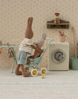 Two plush Maileg rabbits, one pushing the other in a toy stroller, stand in a miniature laundry room with a washing machine, drying rack, potted plants, and Maileg Carrots in Bag for a cute touch to imaginative play.