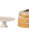 The Maileg Party Cake by Maileg is displayed on a white decorative cake stand beside pancakes with white icing and colorful fruit, all set against a plain background.