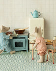 Two Maileg mice in pajamas enjoy their miniature Kitchen set, which features a blue stove, table with chairs and plate, small fridge, and blue teapot—all arranged on a tiled floor for delightful pretend play.