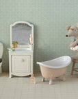 The Maileg Little Sister Beach Mouse by Maileg sits on a stool in a miniature bathroom with a toilet, sink, bathtub, and patterned wallpaper. She wears glasses, a swimsuit, and a swim ring for adorable beach-themed play.