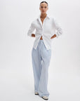 A person stands against a plain white background wearing the Rag & Bone connie shirt, a relaxed-fit cotton style with rolled sleeves, paired with light blue and white striped wide-leg pants and white shoes.