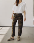 Someone stands indoors by a plain light wall, wearing a loose white tee, beige slip-on shoes, and Hey Gang’s The Officer Pant in 100% Cotton Ripstop with rolled cuffs. Their face is not visible.