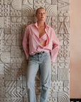 A person stands against a textured wall with geometric patterns wearing relaxed fit light blue jeans and a pink Cissa The Daily Shirt open to reveal a pink bra, looking confidently at the camera.