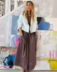 A woman wears the KR Tank Dress by Kerri Rosenthal with a light jacket, leaning against a wall indoors. Large colorful abstract paintings enhance the modern, artistic vibe of the setting.