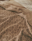A close-up of Coyuchi Inc’s Mirada Matelasse Blanket shows beige and light brown jacquard fabric in subtle floral and geometric patterns, evoking a vintage Indian woodblock look with softly wrinkled, layered texture.