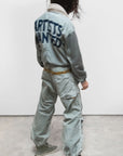A person with wavy dark hair wears Free City's Flap/Snap AirJump Pant in light gray with a custom sunfade wash and "ARTISTS WANTED" on the back, green high-top sneakers, and a white scarf, standing on concrete facing away from the camera.