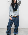 A person poses confidently against a plain background, wearing a loose gray sweatshirt, Free City (sparrow, LLC) Matte/Satin Biggy Pant in an oversized fit, olive green high-top sneakers, a belt, and has dark wavy hair framing their face.