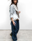 A person with long wavy hair, in a Free City (sparrow, LLC) A. W. Heather Bleachout Sweatshirt, beige scarf, loose blue pants, and green sneakers walks indoors on a concrete floor against a plain white wall.