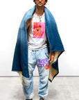 A man stands against a plain background, wearing a dark cap, white shirt with “THE FORCE HAS A LOT OF POWER” in orange and pink, light blue FREE CITY sweatpants, green sneakers, and the Free City Indigo Sherpa Blanket/Indigo Blues.