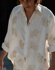 A person wears The Mr Big Shirt by Cissa—a loose cotton shirt with a beige floral pattern and adjustable cuffs. Sleeves are rolled up and only the upper body is visible, set against a softly blurred background.