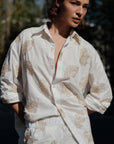 A person stands outdoors in the Cissa Mr Big Shirt—a loose, white cotton shirt with adjustable cuffs—and matching beige floral pants, hands in pockets as sunlight highlights their face and upper body.