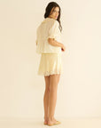 A woman with long dark hair stands sideways, barefoot, against a plain light background, wearing the light yellow DOEN Henri Top with short sleeves and lace trim, paired with a matching skirt.