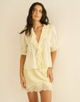A woman with long brown hair wears the DOEN Henri Top—a cream lace ruffle shirt with puffed sleeves—and a matching cream mini skirt with lace trim, standing against a beige background.