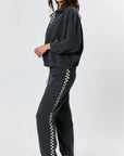 A person stands sideways in Ragabond Studio’s Studio Sweats Checkerboard, featuring a black-and-white checkerboard stripe, paired with black shoes and white socks against a plain light background.