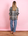 A person with long blonde hair stands facing away, wearing the KUJTEN Lumber L/S Shirt in gray plaid, blue jeans, and sandals, set against a pink background and wooden floor.