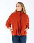 A woman stands against a white background, wearing the Kerri Rosenthal Coze Magoze cotton fleece jacket with blue trim and snap buttons, paired with dark blue jeans. She looks to the side, hands in her jacket pockets.