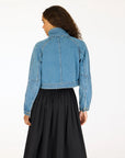 A woman with long, wavy hair faces away, showcasing the back of a Kerri Rosenthal Denim Jacket with heart elbow patches and a high collar, paired with a black skirt against a plain background.