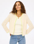 A woman with long curly hair wears the Frankie Beau-Lero open front cotton crochet cardigan by Kerri Rosenthal, featuring light yellow trim and subtle heart detail, over a cream top and blue jeans, posing against a plain white background.