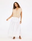 A woman poses against a white background wearing Kerri Rosenthal's Vacance Pintuck Skirt—a long white poplin skirt with tiered lace trim—paired with a beige short-sleeve button-up, brown sandals, and a gold watch for an effortlessly elegant look.