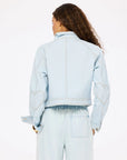 A person with long, curly hair models the Kerri Rosenthal Denim Jacket—light blue, double breasted, with heart elbow patches—and matching denim pants. They are shown from behind against a plain background.
