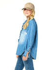 A person in a blue denim shirt and jeans stands sideways, smiling. Their long hair flows from beneath a beige hat, effortlessly stylish against a plain white background, reminiscent of the KR Imperfect Heart Hat by Kerri Rosenthal.