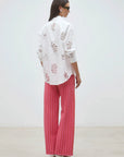 A woman with long dark hair, sunglasses, and white heels stands in a minimal room wearing the SUNCOO Loula Blouse—an embroidered white 100% organic cotton button-down—and loose pink checkered pants. She is facing away.