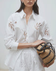 A woman wears the SUNCOO Loula Blouse, a white organic cotton button-up with cut-out details, paired with a matching skirt and holding a woven straw bag with black handles against a plain light background.