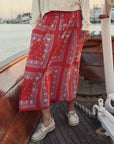 Wearing The Canary Skirt by The Great Inc., featuring a red pattern, and beige boat shoes, a person stands on a wooden boat with water and docked sailboats in the background.