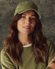 A woman with long brown hair wears The Classic Baseball Hat by The Great Inc., featuring vintage embroidery and an adjustable back, paired with a green sweater over a white shirt, standing before a neutral textured background, smiling slightly at the camera.