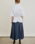 A person stands facing away, wearing the Aquarius Cocktail Le Cargo Skirt in blue plaid, a loose white Japanese cotton poplin shirt cinched with a belt, black argyle socks, and heels. Their hair is tied back against a neutral background.