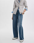 A person wearing a light gray plaid blazer, blue striped shirt, wide-leg blue jeans with side detailing, and Rag & Bone’s lexi loafers stands with one hand in their pocket against a plain background.