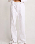 A person stands against a plain light background wearing Denimist’s DECK PANT WHITE WASH, loose-fitting white 100% cotton denim jeans with front pockets. Their face is not visible; they pair the pants with a white partially-tucked shirt and sandals.