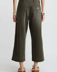 A person faces away, wearing Denimist's olive green 100% cotton Georgia Work Pant with a white top, brown moccasins, and bracelets on both wrists, set against a plain light background.