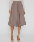A person wears the BROCHU WALKER LOTI BUBBLE SKIRT in taupe—knee-length with a fitted waistband—paired with a light long-sleeve top and beige pointed-toe heels, standing against a plain background.