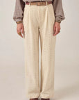 A person wears Maison Hotel's Theodore Pant—high-waisted, wide-leg, cream eyelet-embroidered cotton twill—styled with a tucked-in plaid shirt, beige belt, and brown shoes. Only the body from the shoulders down is visible against a neutral background.