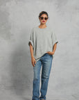 A woman poses before a gray textured backdrop, wearing sunglasses, blue jeans, red flip-flops, and the Mamene Boyfriend Tee—a loose, lightweight knit gray top. Her hair is tied up and one hand rests in her pocket.