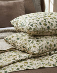 Two Amity Home Millie Euro Shams with green and beige floral pillowcases are stacked on a hand-quilted cotton voile quilt, placed over brown and striped bedding in a softly lit room.