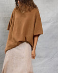 A person wearing Mamene's Boyfriend Tee, a loose brown lightweight knit short-sleeve sweater, with a long beige skirt stands against a light gray textured background, wavy hair down and one hand by their side.