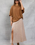 A woman in wavy hair and sunglasses poses against a textured gray background, wearing the Mamene Boyfriend Tee, a loose brown lightweight knit top, paired with a light beige thigh-high slit maxi skirt and green platform sandals.