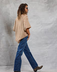 Wearing Mamene's Boyfriend Tee, a person with wavy hair, sunglasses, blue jeans, and brown boots walks before a textured gray backdrop.
