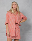 A woman with long blonde hair wears the Mamene Repeat Shirt—a loose-fitting, pink and white Japanese yarn-dye stripe cotton button-down—paired with matching shorts, standing against a light gray textured background, one hand on her hip.