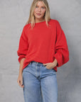 A woman with long blonde hair models the Mamene Winona Sweater, a bright red cotton knit, paired with light blue jeans. She stands against a light gray background, one hand in her pocket, looking at the camera with a slight smile.