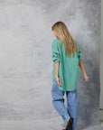 A person with long blonde hair, barefoot and facing away from the camera, models Mamene’s Repeat Shirt—a loose green striped cotton poplin top—paired with blue jeans against a gray textured backdrop.