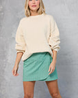 A woman with blonde hair stands against a gray textured background, wearing Mamene's Repeat Wrap Tennis Skirt in green Japanese yarn-dye poplin with white stripes and a loose cream sweater. She looks relaxed and confident.