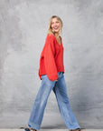 A smiling woman with blonde hair glances left as she walks in front of a gray textured backdrop, wearing the Mamene Winona Sweater in red, paired with blue jeans and flip-flops.