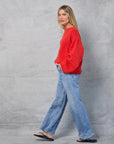 A woman with long blonde hair wears the Mamene Winona Sweater, an oversized bright red knit, paired with wide-leg blue jeans and flip-flops as she walks in front of a gray textured background.