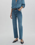 A person wears a tucked-in denim shirt, high-waisted blue jeans with a black belt, and Rag & Bone's mercer slingback flat—sleek black leather shoes with cushioned footbed—one hand in pocket, standing against a plain background.