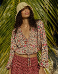 Wearing the natalie martin Amelia Top, a person stands before large palm leaves. Sunlight casts shadows on their cropped long-sleeve v-neck top, patterned shorts, and beige bucket hat.